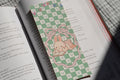 Cute linen bookmark featuring an adorable bunny reading a book on a pastel green checkerboard background tucked inside an open book.
