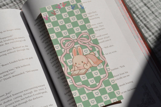 Cute linen bookmark featuring an adorable bunny reading a book on a pastel green checkerboard background tucked inside an open book.
