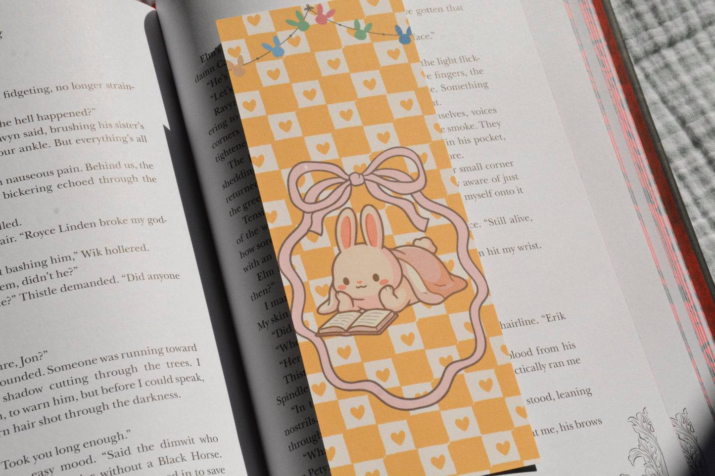 Pastel linen bookmark featuring a cozy reading bunny illustration placed between the pages of a novel.
