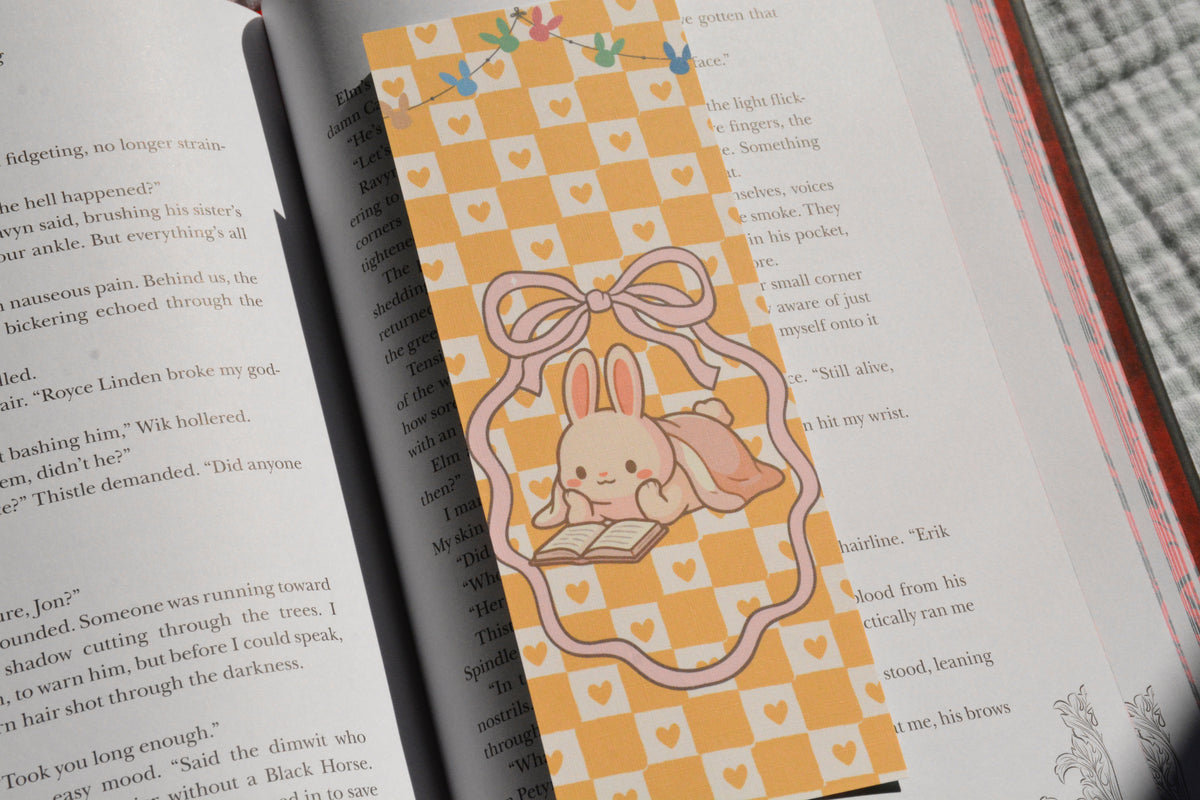 Pastel linen bookmark featuring a cozy reading bunny illustration placed between the pages of a novel.
