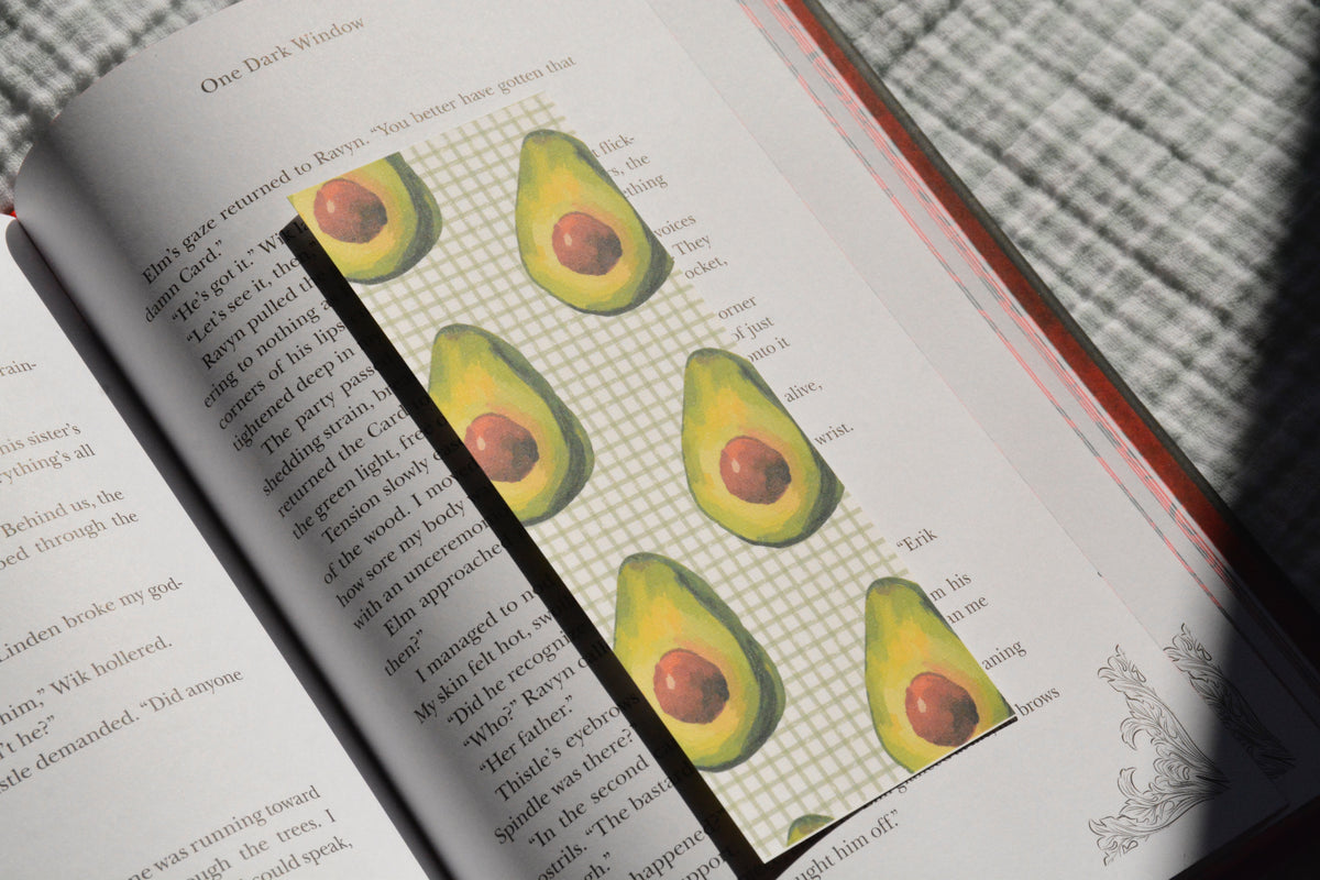 Handcrafted linen bookmark featuring a watercolor avocado pattern resting inside an open book.
