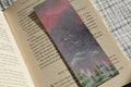 Double-sided linen bookmark featuring a dreamy moonlit forest on one side and a warm sunrise landscape with evergreen trees on the other.