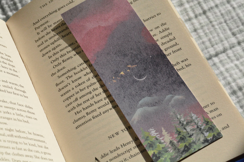 Double-sided linen bookmark featuring a dreamy moonlit forest on one side and a warm sunrise landscape with evergreen trees on the other.