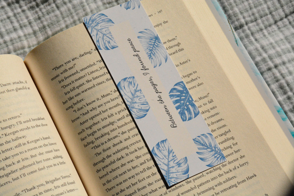 Linen bookmark featuring blue botanical leaves and the quote "Between the pages, I found peace" placed inside an open book.