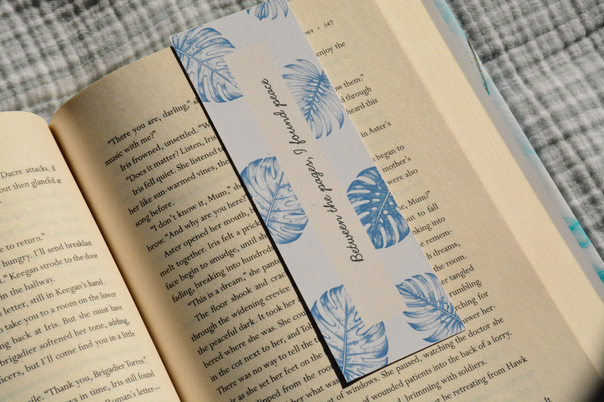Linen bookmark featuring blue botanical leaves and the quote "Between the pages, I found peace" placed inside an open book.
