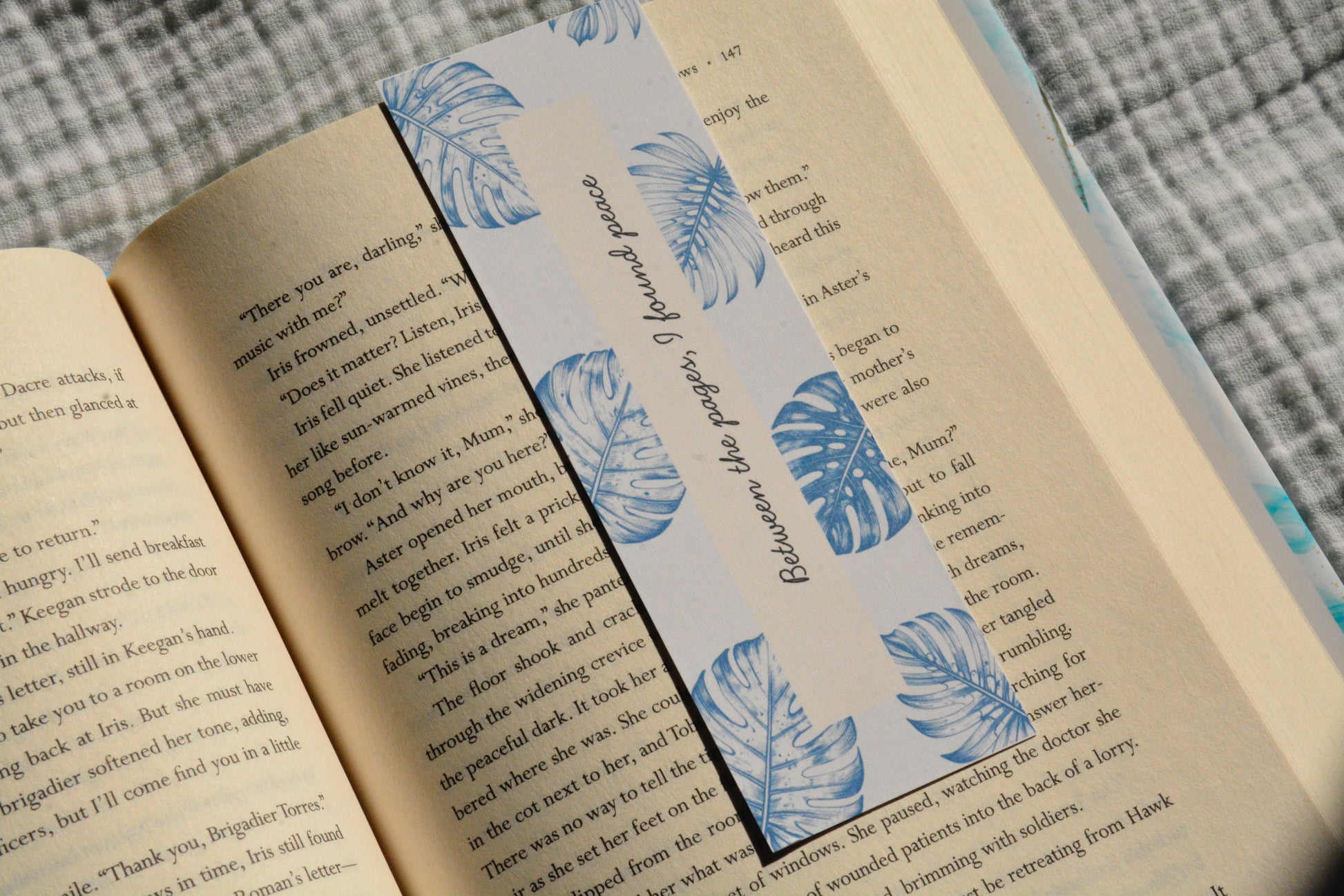 Linen bookmark featuring blue botanical leaves and the quote "Between the pages, I found peace" placed inside an open book.