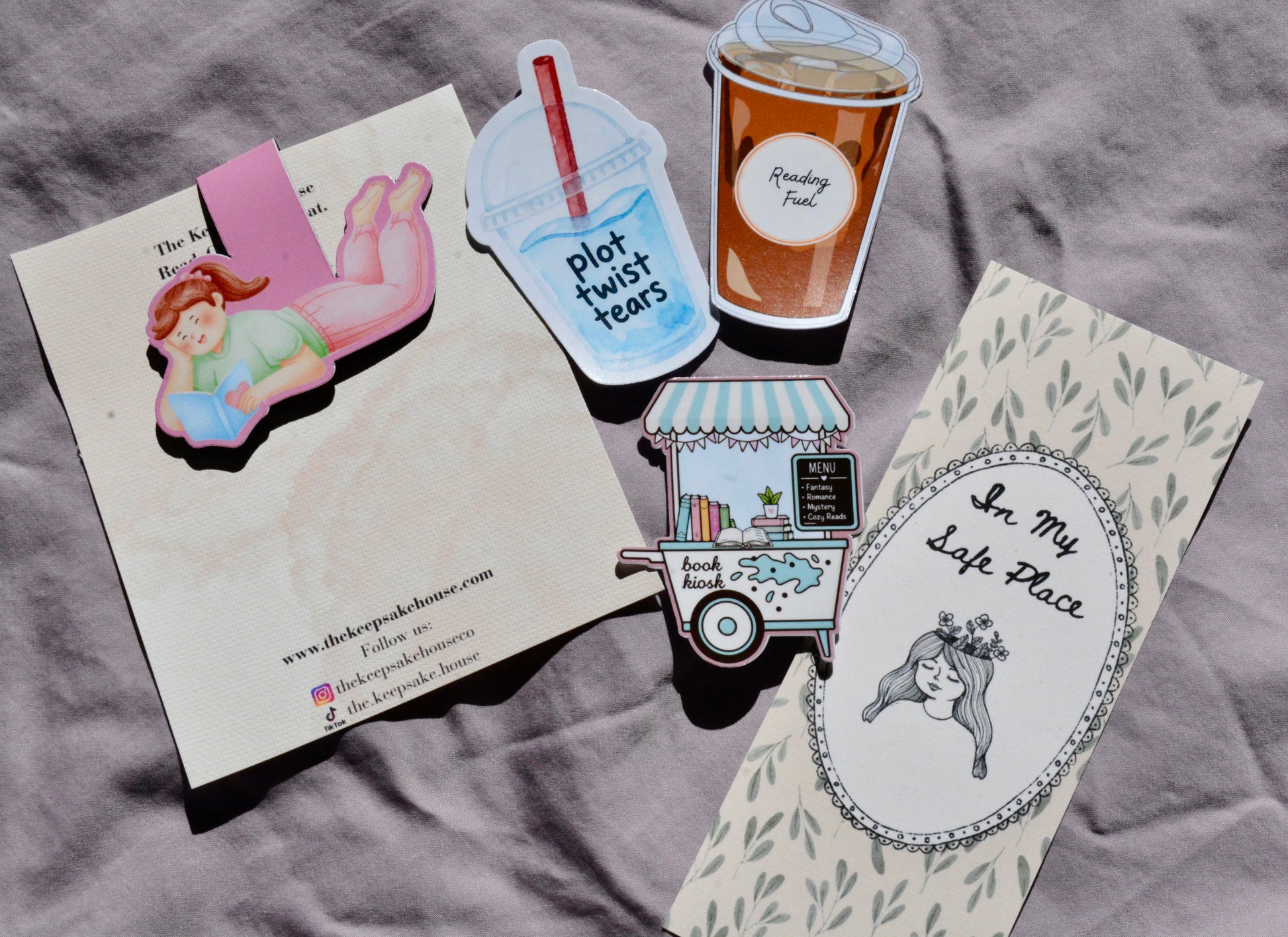A flat-lay of several printed paper items on a gray fabric background, including a cream card with a pink illustrated character, a blue drink cup sticker reading 'plot twist teas,' a brown coffee cup sticker, a small food cart sticker, and a cream oval pin or patch with the text 'In My Left Place' and a black-haired illustrated woman.