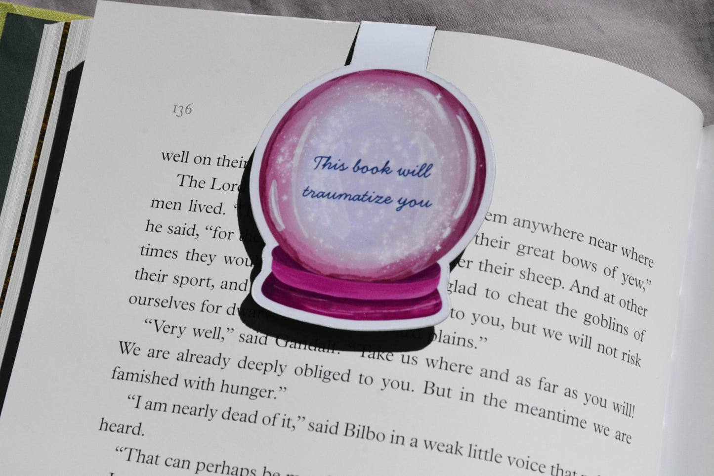 This Book Will Traumatize You magnetic bookmark clipped inside an open fantasy novel.