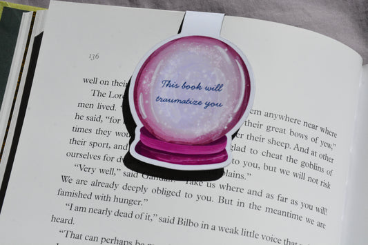 This Book Will Traumatize You magnetic bookmark clipped inside an open fantasy novel.
