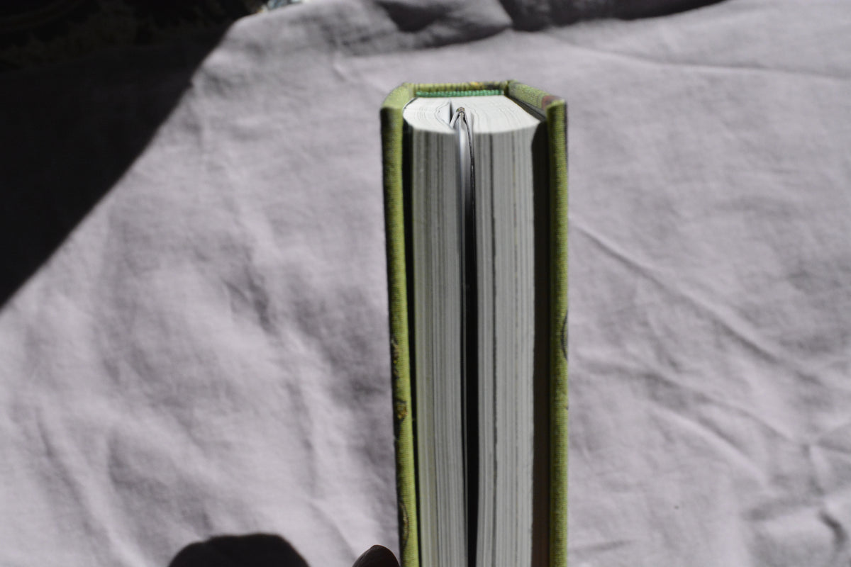 A close-up of a book standing upright, showing its green cloth binding and white pages, photographed against a wrinkled light gray fabric background.