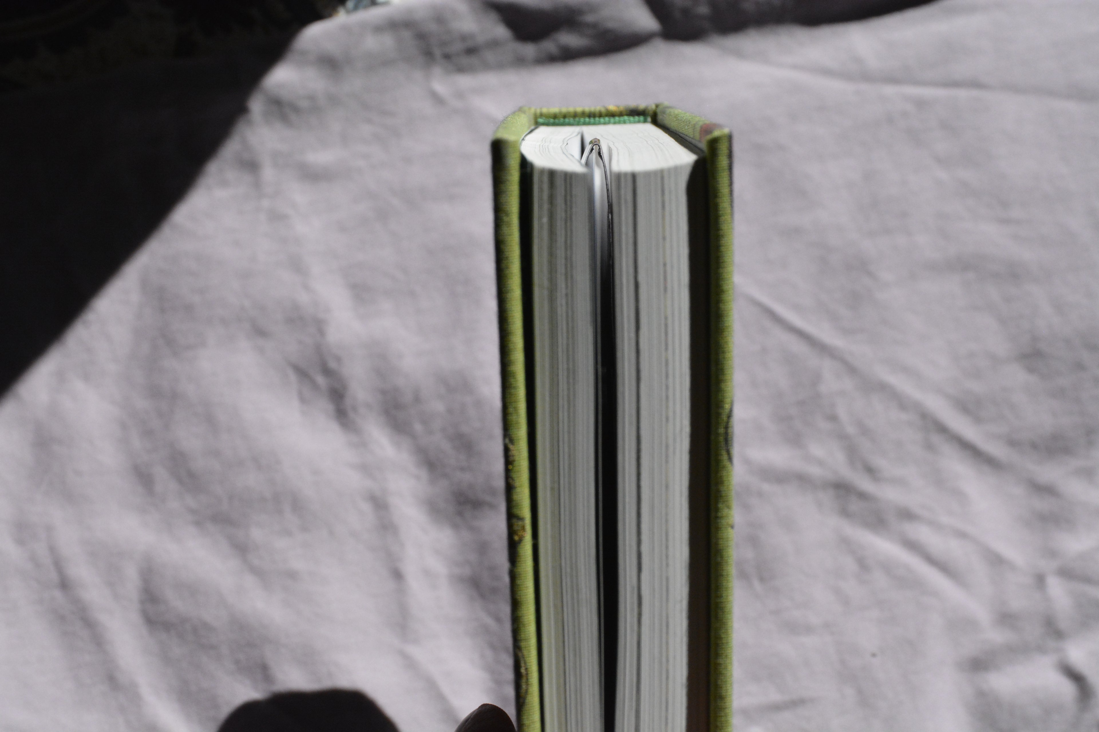 A close-up of a book standing upright, showing its green cloth binding and white pages, photographed against a wrinkled light gray fabric background.