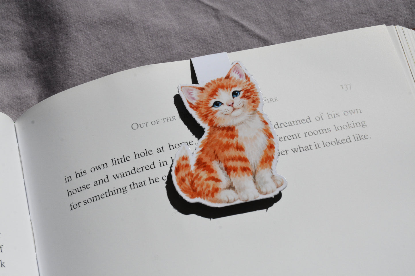 A magnetic bookmark of an orange and white tabby kitten sitting on an open book page, with a gray fabric surface visible in the background.