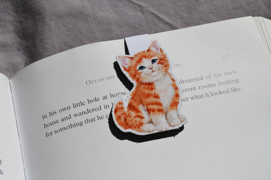 A magnetic bookmark of an orange and white tabby kitten sitting on an open book page, with a gray fabric surface visible in the background.