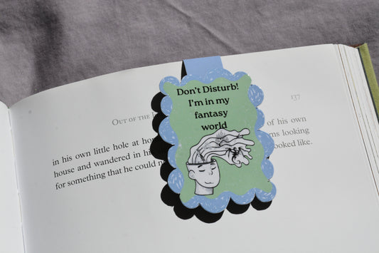 A magnetic bookmark attached to an open book page. The bookmark has a light blue top and bottom border, a pale green center, black scalloped edges, and a cartoon illustration of a hand reaching toward a brain with the text 'Don't Disturb! I'm in my fantasy world.
