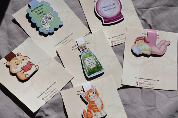 Six illustrated magnetic bookmark on a gray fabric background. The designs include a hamster reading, a blue label with a rabbit, a purple potion bottle, a pink fairy tale girl, a green potion bottle, and an orange tabby kitten.