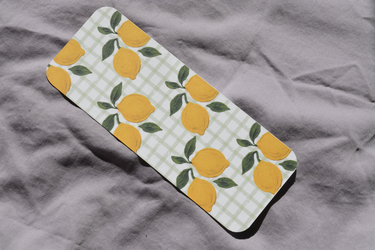 A rectangular mat with rounded corners featuring a yellow lemon and green leaf pattern over a white gingham background, placed diagonally on a textured gray fabric surface.