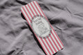 A rectangular book or notebook with pink and white vertical stripes, featuring a central white oval label with the words 'Never Say Reading' and a black line drawing of a woman reading, placed on a wrinkled gray fabric background.
