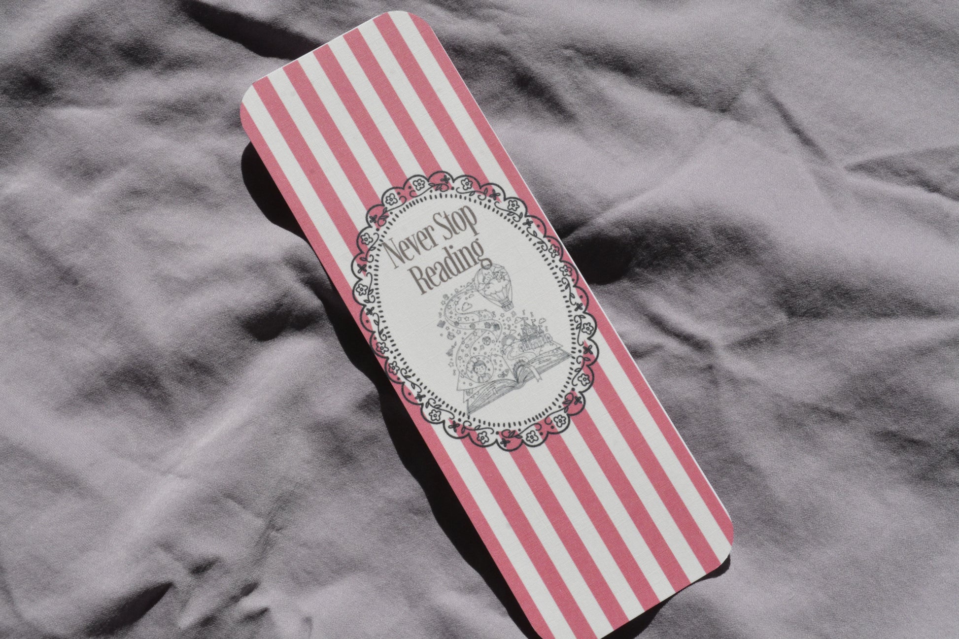 A rectangular book or notebook with pink and white vertical stripes, featuring a central white oval label with the words 'Never Say Reading' and a black line drawing of a woman reading, placed on a wrinkled gray fabric background.