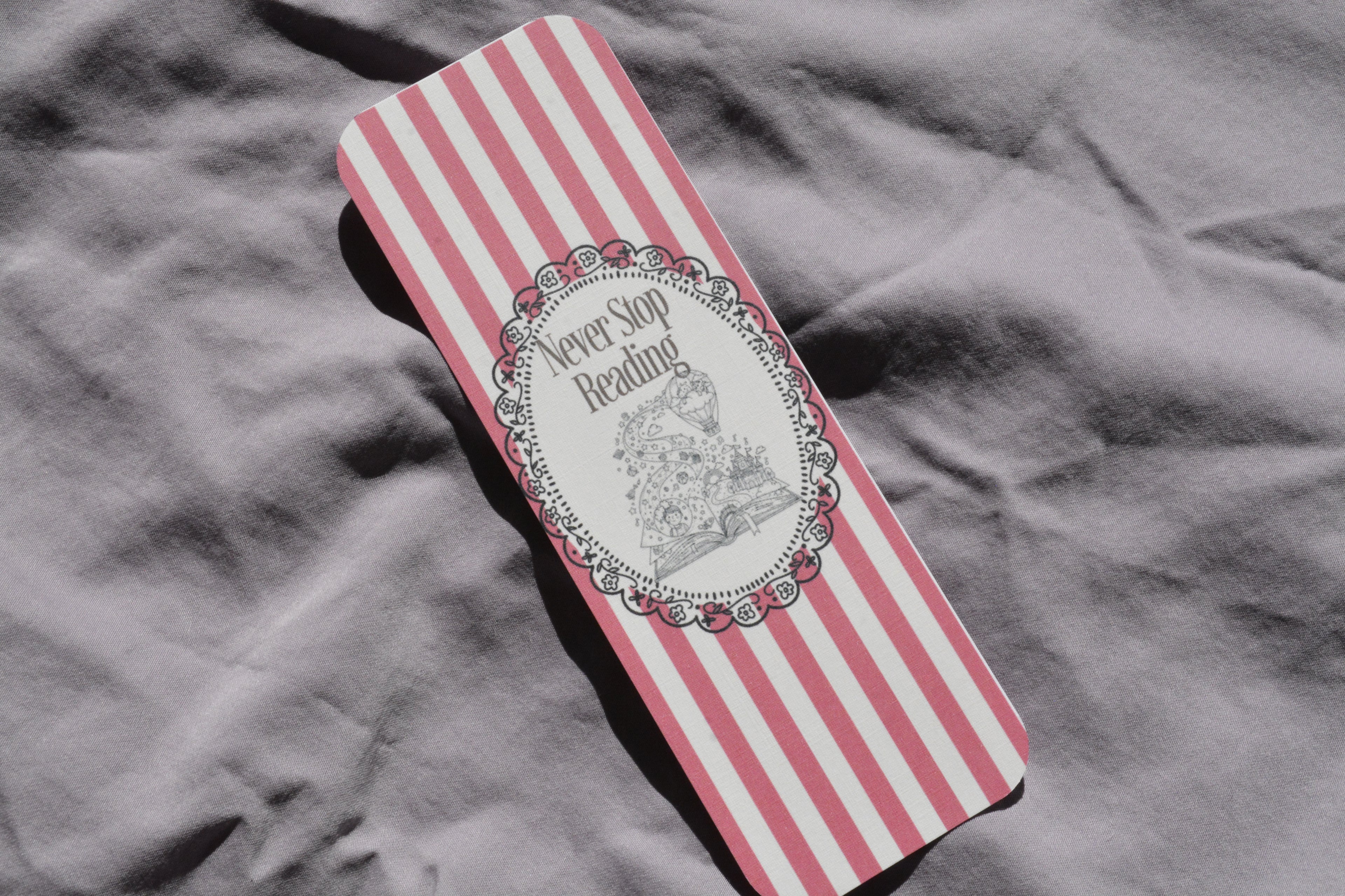 A rectangular book or notebook with pink and white vertical stripes, featuring a central white oval label with the words 'Never Say Reading' and a black line drawing of a woman reading, placed on a wrinkled gray fabric background.