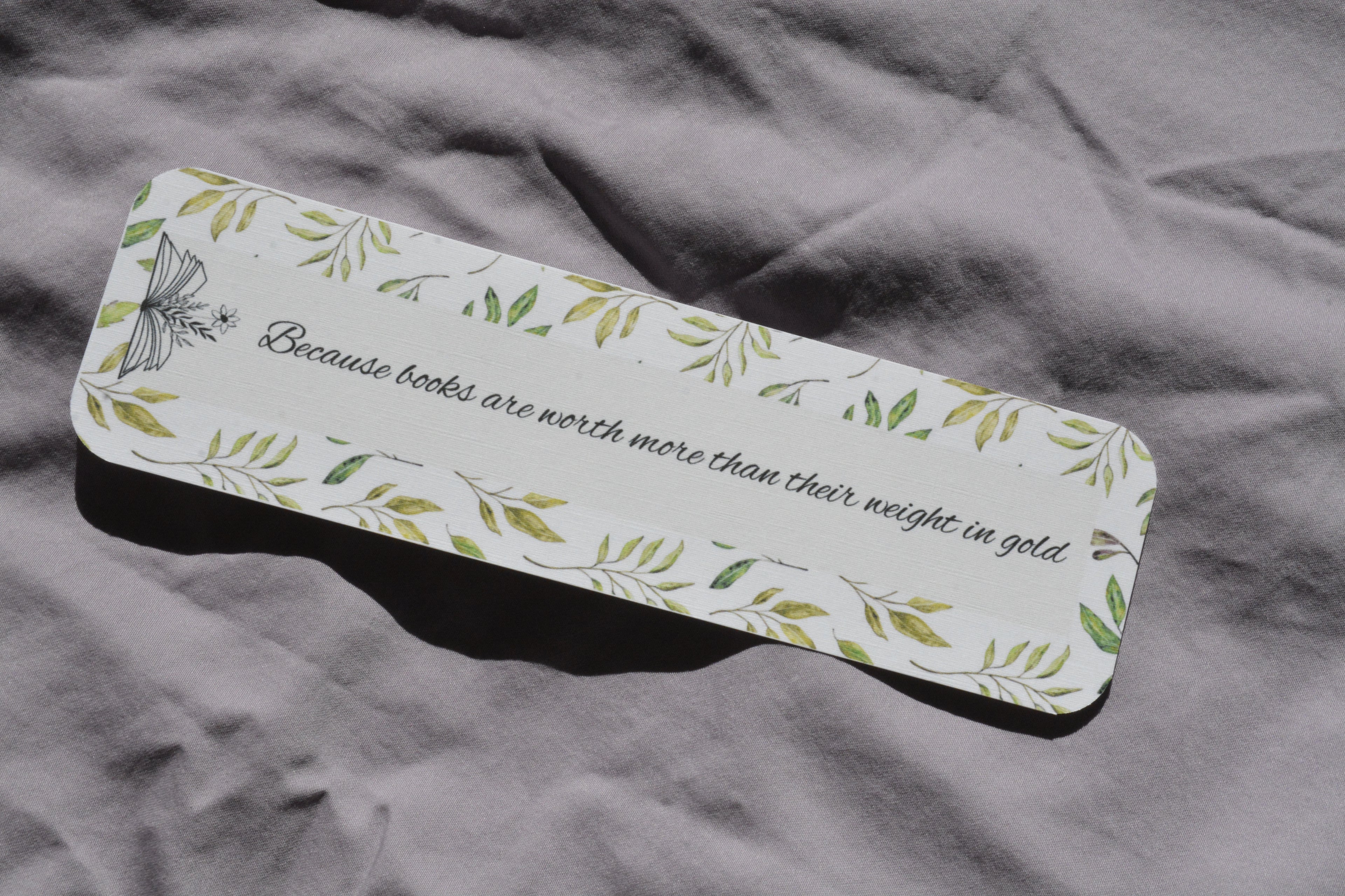 Rounded corner linen bookmark with watercolor leaves and the quote "Because books are worth more than their weight in gold" inside an open book.