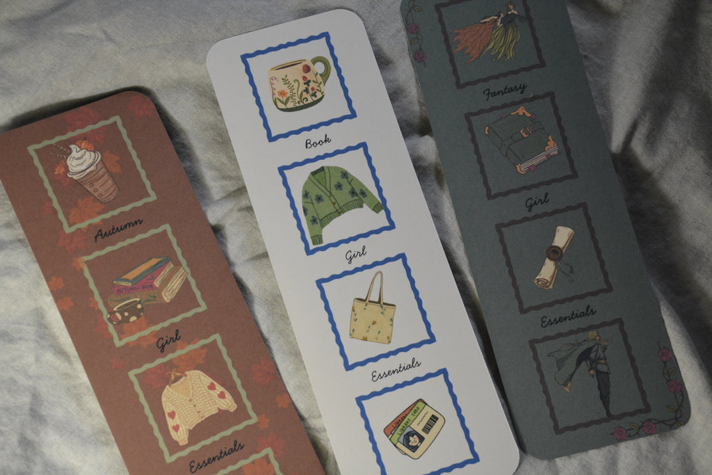 Three illustrated metal bookmarks laid on a light gray fabric background. One bookmark is brown with autumn-themed illustrations and labels; one is white with blue borders and central label; one is dark gray with muted illustrations and labels.
