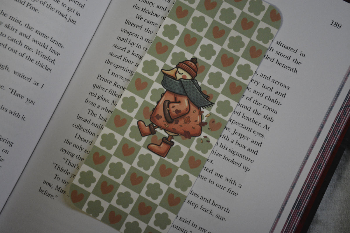 A decorative book bookmark shaped like a child in a winter outfit walking on a green patterned background with small red hearts, placed on an open book.