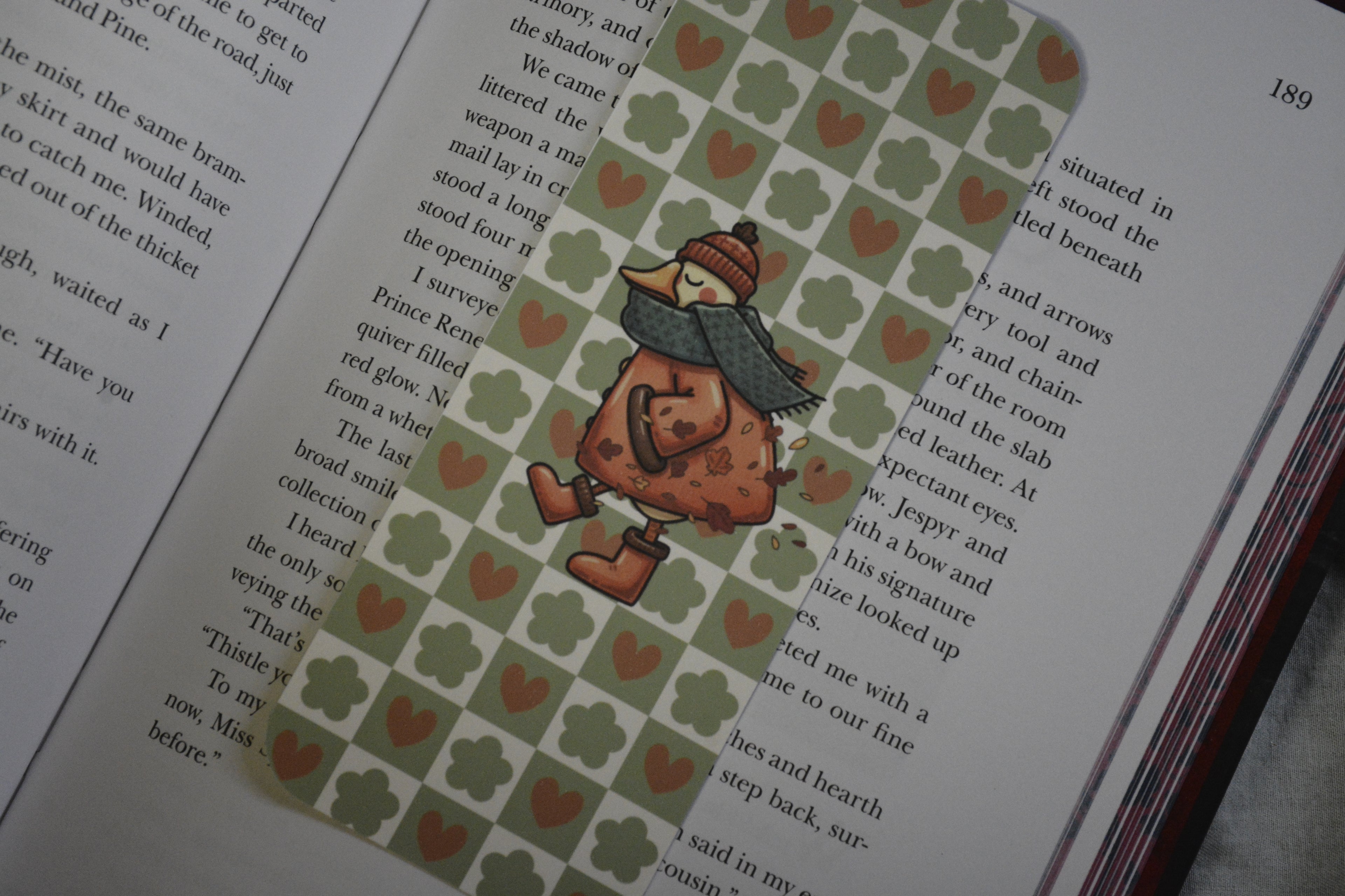 A decorative book bookmark shaped like a child in a winter outfit walking on a green patterned background with small red hearts, placed on an open book.
