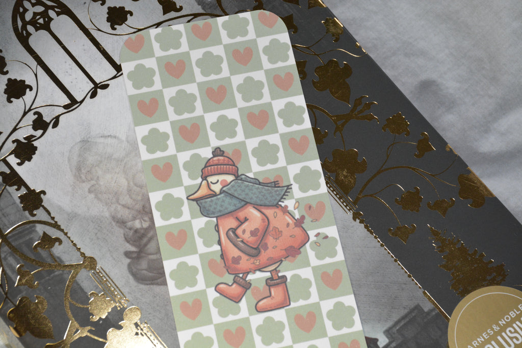 A rectangular printed card or bookmark featuring a whimsical duck character in a winter outfit, placed diagonally over a gray patterned sheet with gold foil floral details.