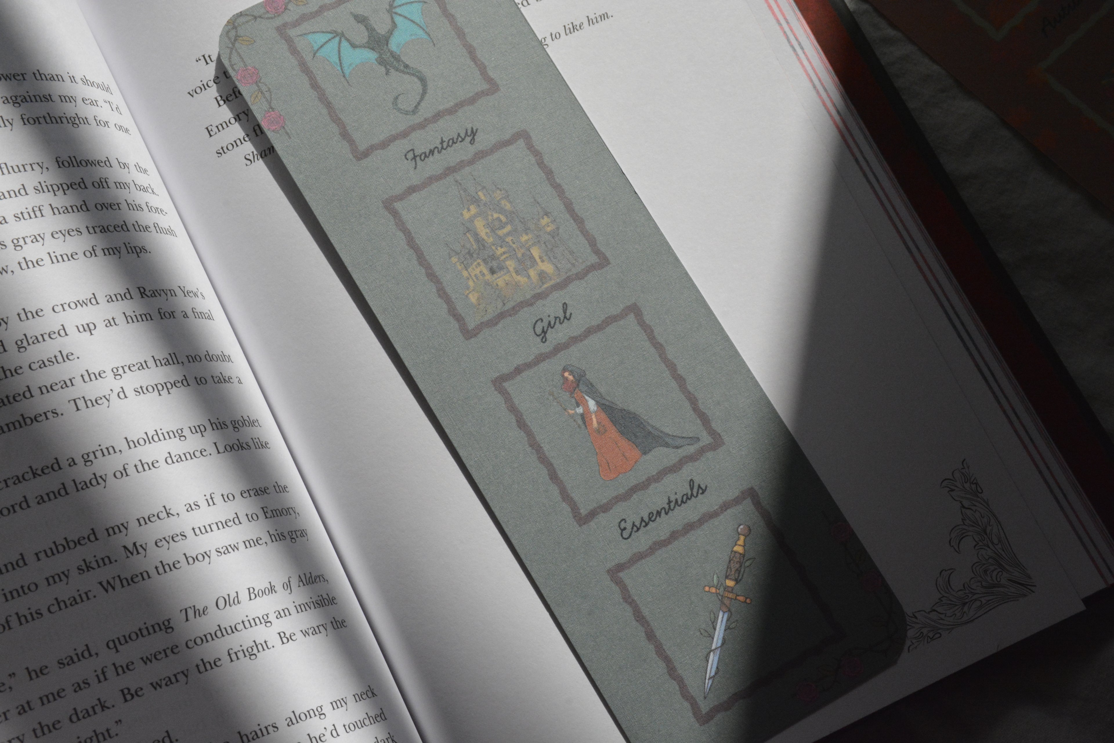 A gray book bookmark with fantasy-themed illustrations, shown resting on an open book.