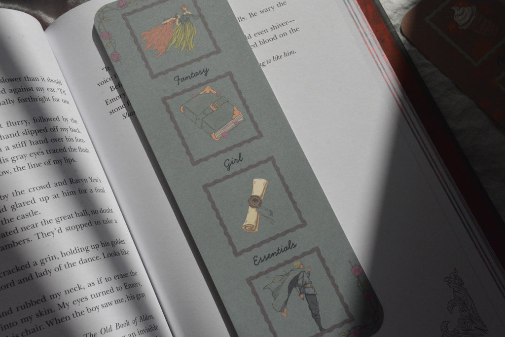 A gray bookmark with illustrated book genres (Fantasy, Sci-Fi, Girl, Essentials) resting on open book pages.