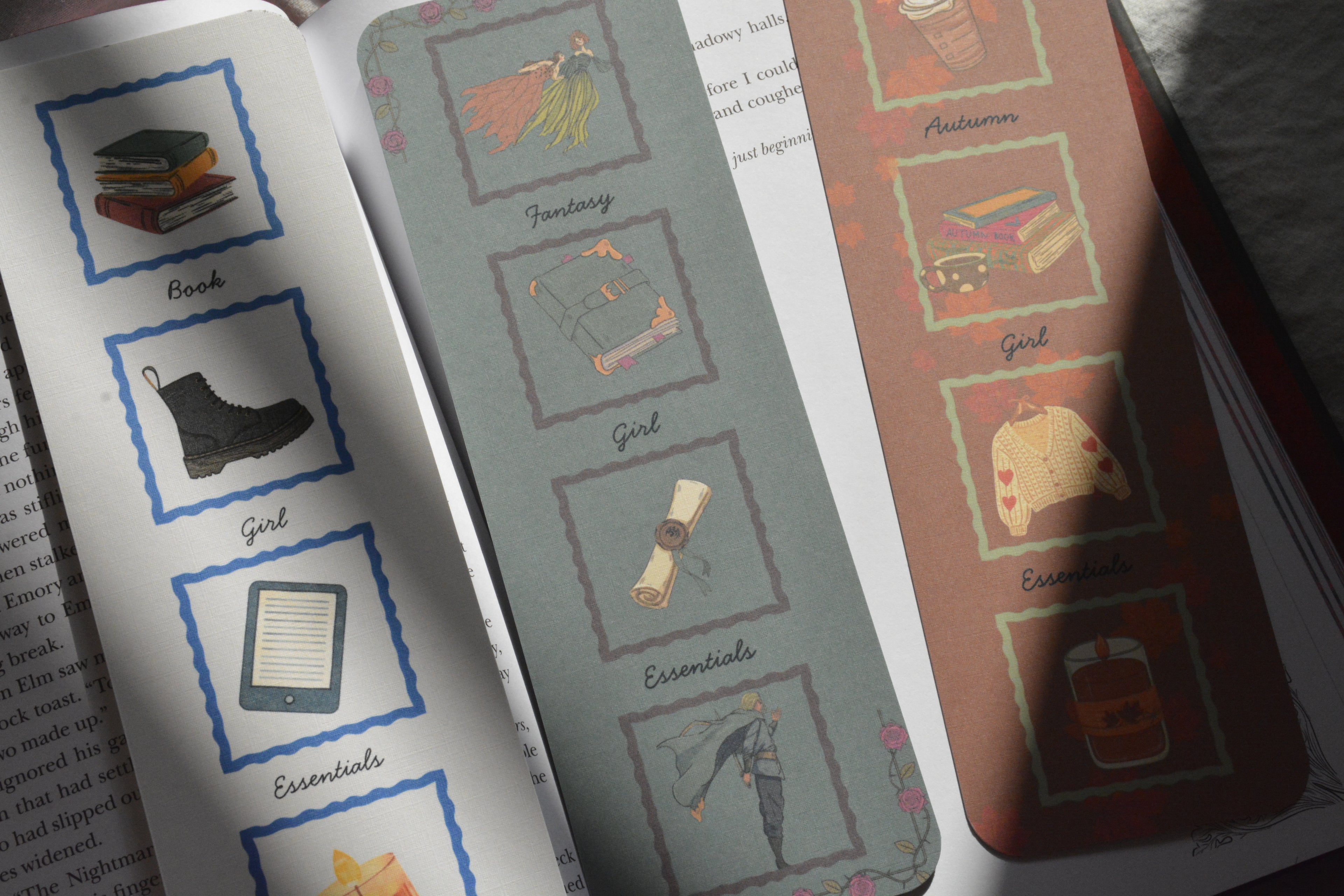Three decorative printed bookmarks or tabs laid over an open book. The bookmarks feature illustrated categories such as Book, Boot, Girl, Essentials, Fantasy, Girl, and Autumn with small drawings inside bordered boxes.