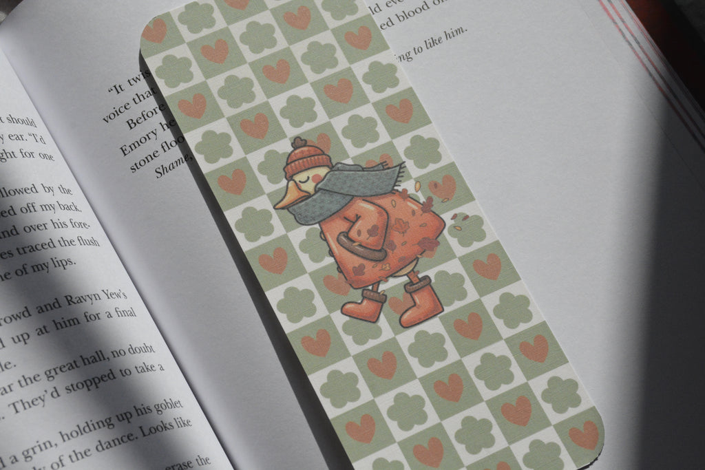 A close-up of an open book with a decorative bookmark featuring a pattern of sage green clovers and pink hearts, with a cartoon duck character wearing a red hat and gray scarf.