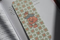 A close-up of an open book with a decorative bookmark featuring a pattern of sage green clovers and pink hearts, with a cartoon duck character wearing a red hat and gray scarf.