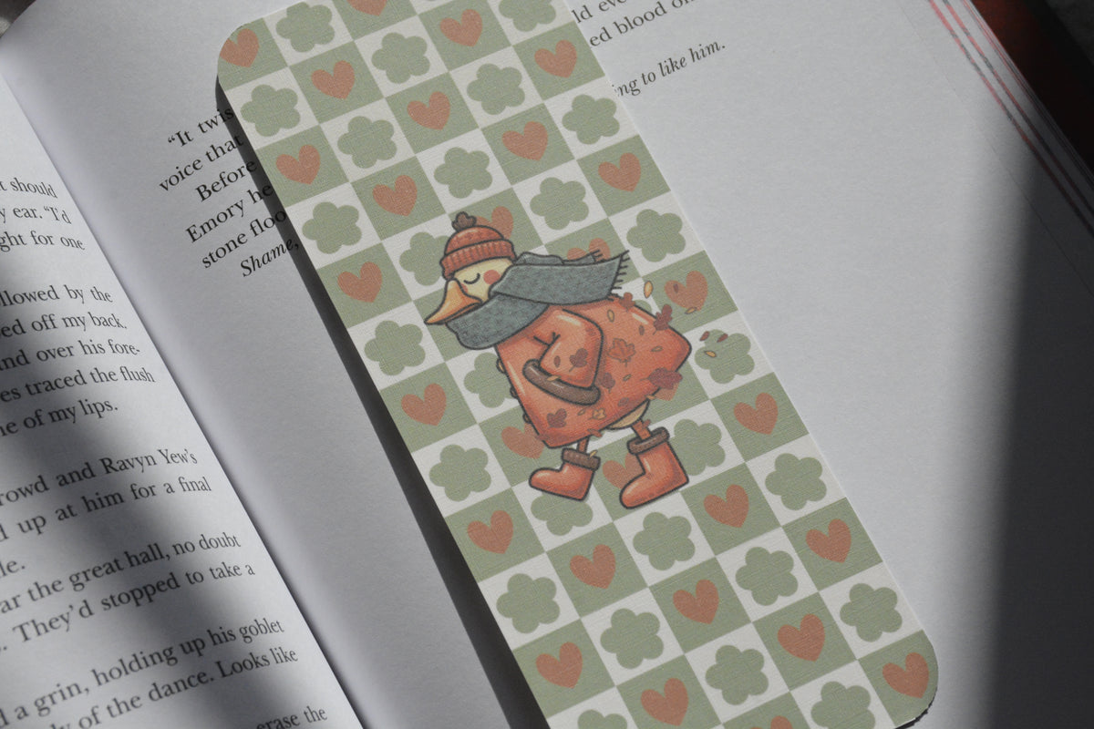 A close-up of an open book with a decorative bookmark featuring a pattern of sage green clovers and pink hearts, with a cartoon duck character wearing a red hat and gray scarf.