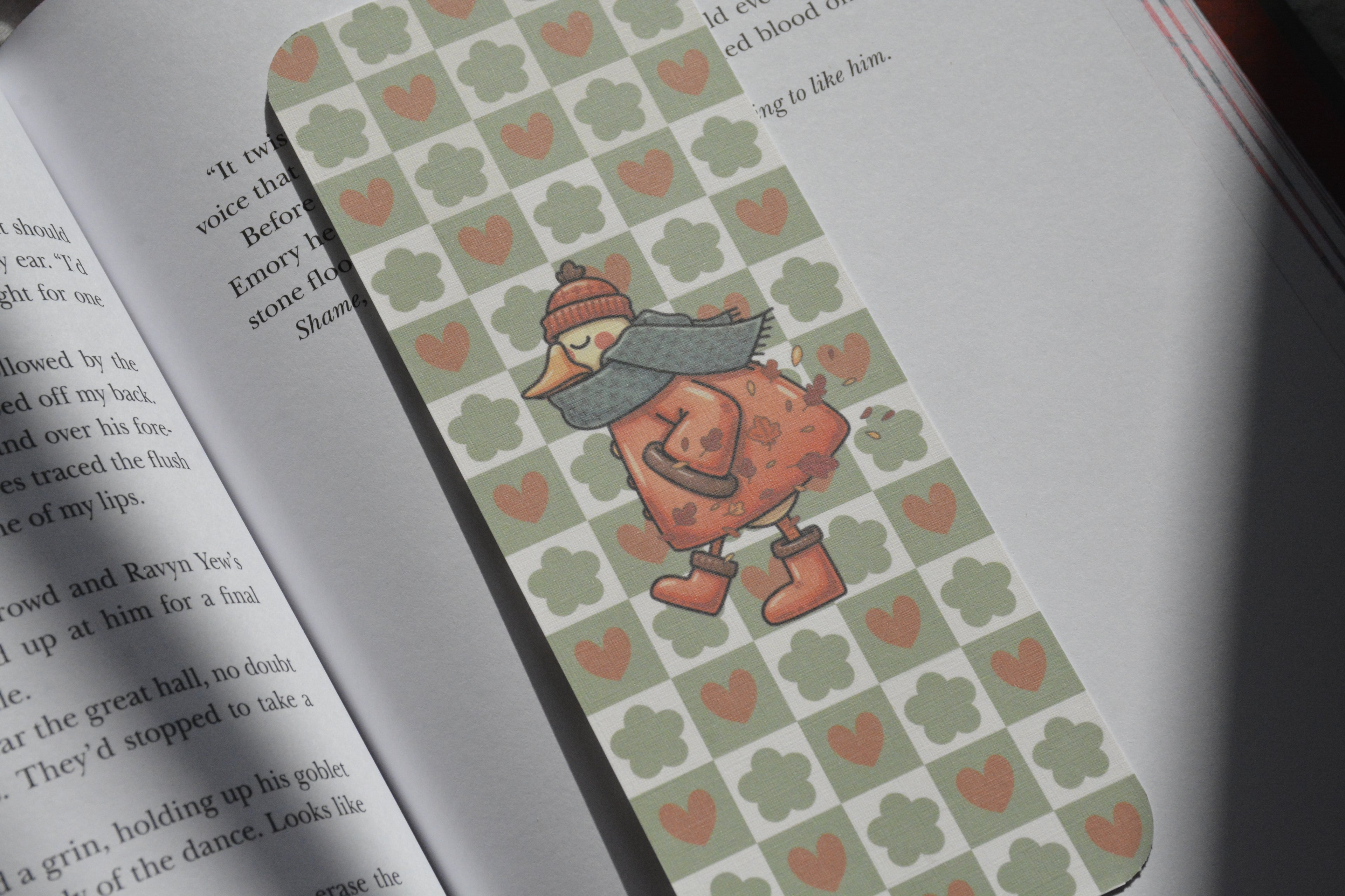 A close-up of an open book with a decorative bookmark featuring a pattern of sage green clovers and pink hearts, with a cartoon duck character wearing a red hat and gray scarf.