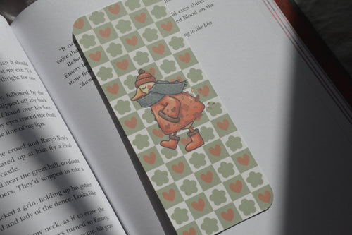 A rectangular book bookmark with a green and orange heart pattern and a cartoon duck character, placed on an open book.
