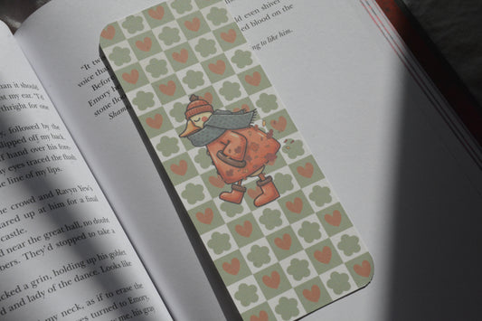 A rectangular book bookmark with a green and orange heart pattern and a cartoon duck character, placed on an open book.