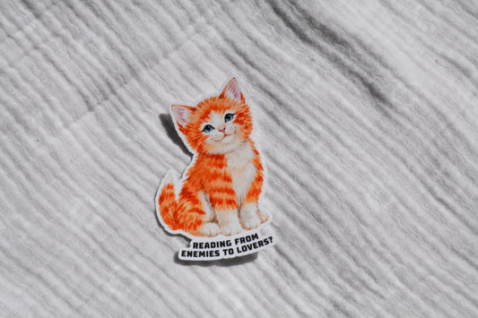 An orange and white tabby kitten sticker with the text 'READING FROM ENEMIES TO LOVERS?' lies on a textured white fabric background.
