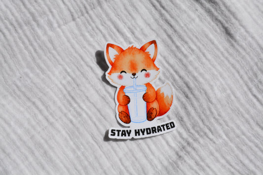 A die-cut sticker featuring a cute orange cartoon fox drinking from a light blue tumbler, with the text 'STAY HYDRATED' below, placed on a white textured fabric background.