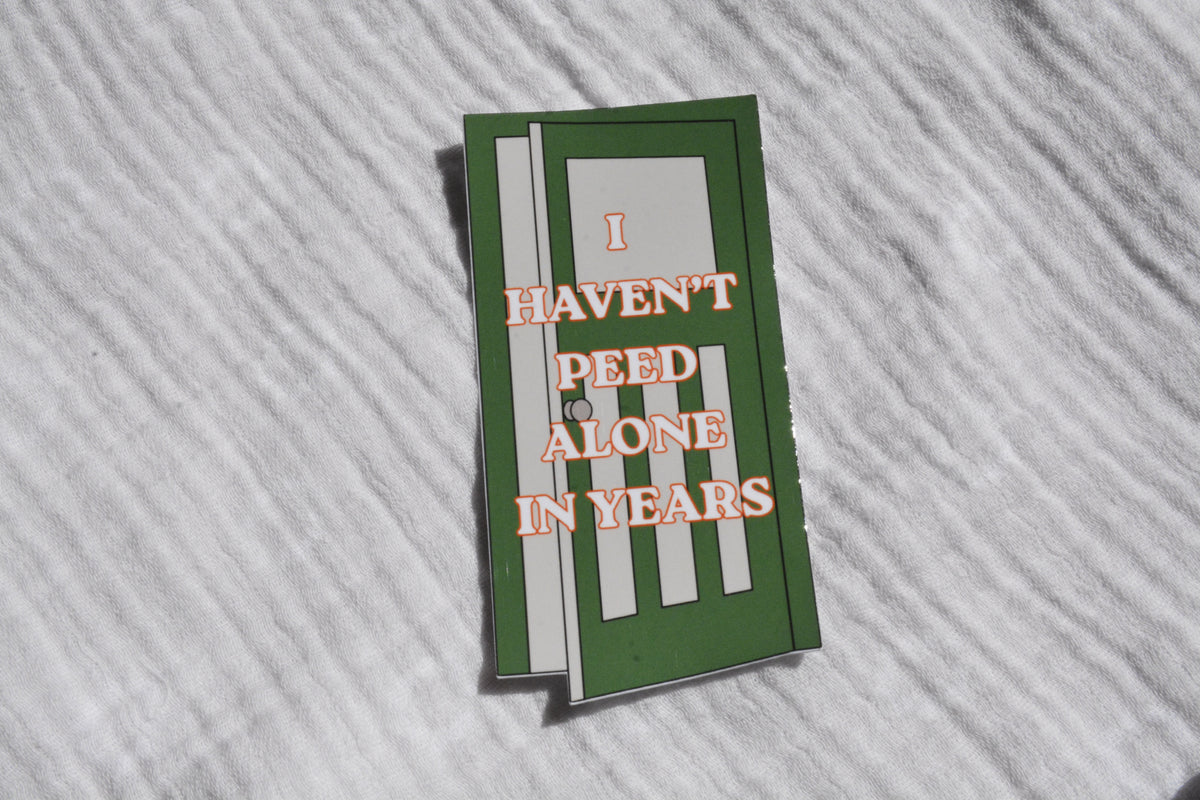 A rectangular sticker with a green background and white stripes, featuring the text 'I HAVEN'T PEED ALONE IN YEARS' in orange outline lettering, displayed on a white textured fabric background.