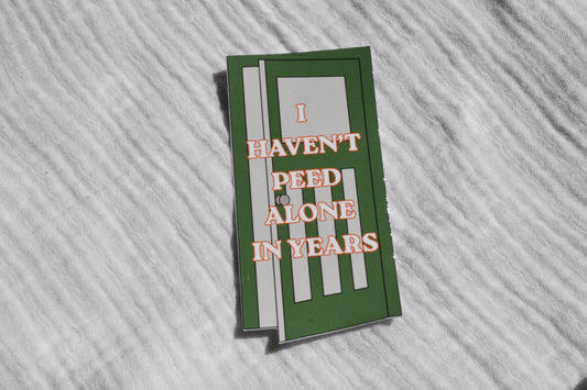 A rectangular sticker with a green background and white stripes, featuring the text 'I HAVEN'T PEED ALONE IN YEARS' in orange outline lettering, displayed on a white textured fabric background.