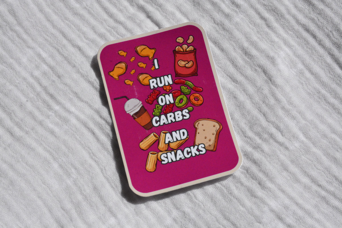 A rectangular sticker with rounded corners featuring a bright pink background and cartoon snack illustrations, placed on a white textured fabric surface. The sticker reads 'I RUN ON CARBS AND SNACKS' with chips, a drink, fruit, and bread graphics.