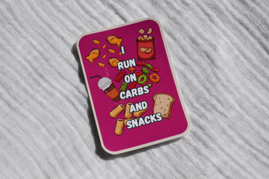 A rectangular sticker with rounded corners featuring a bright pink background and cartoon snack illustrations, placed on a white textured fabric surface. The sticker reads 'I RUN ON CARBS AND SNACKS' with chips, a drink, fruit, and bread graphics.