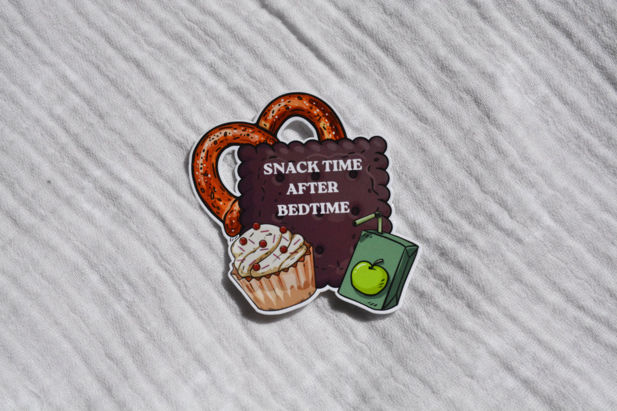 A die-cut sticker featuring a pretzel, a cupcake, and a juice box with the text 'SNACK TIME AFTER BEDTIME' on a white textured fabric background.