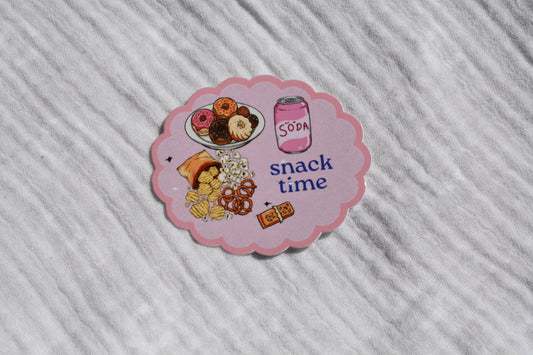 A scalloped pink sticker featuring cartoon illustrations of donuts, a soda can labeled 'SODA', and snacks with the text 'snack time', placed on a textured white fabric background.
