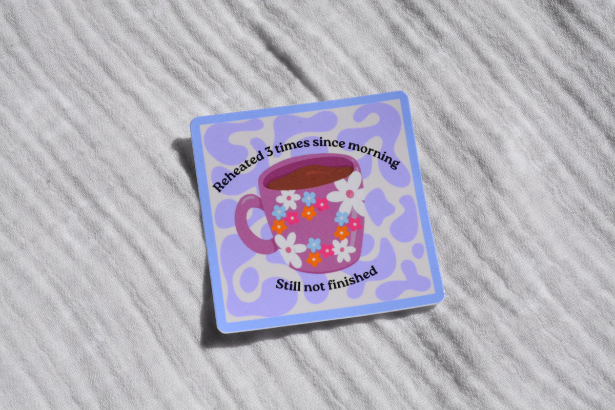 A square sticker with rounded corners featuring a purple coffee cup with white, pink, and blue flowers, surrounded by a light blue border. The sticker has black text reading 'Reheated 3 times since morning' and 'Still not finished'.