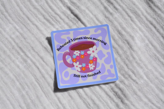 A square sticker with rounded corners featuring a purple coffee cup with white, pink, and blue flowers, surrounded by a light blue border. The sticker has black text reading 'Reheated 3 times since morning' and 'Still not finished'.