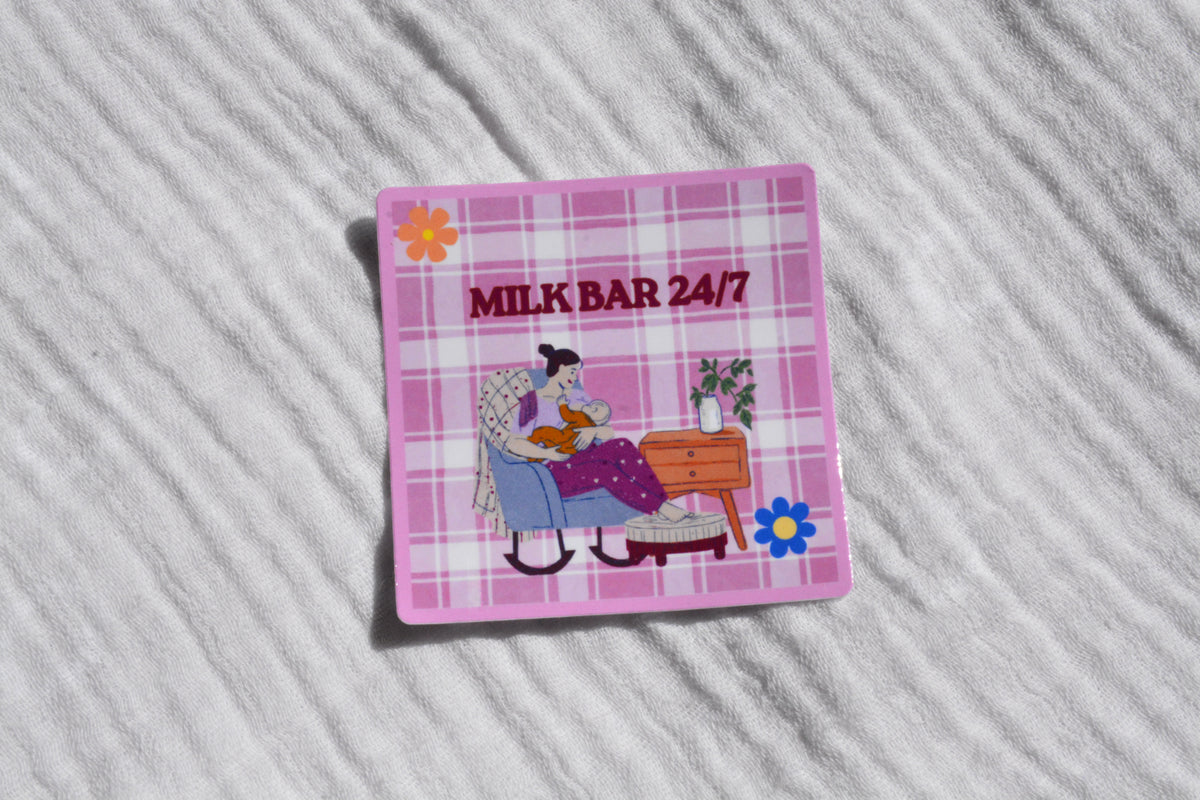 A square pink plaid sticker featuring the text 'MILK BAR 24/7' above an illustration of a woman reading in a chair, placed on a white textured fabric background.