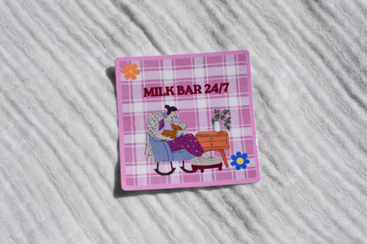 A square pink plaid sticker featuring the text 'MILK BAR 24/7' above an illustration of a woman reading in a chair, placed on a white textured fabric background.