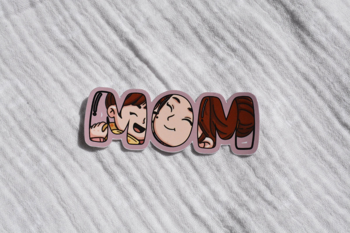 A die-cut sticker spelling 'MOM' in stylized cartoon letters, placed on a textured white fabric background.